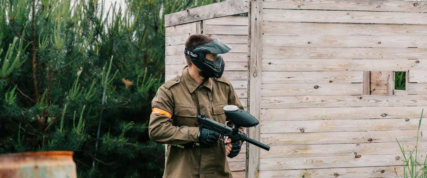 Gracz paintballowy w mundurze na tle drewnianej przeszkody.